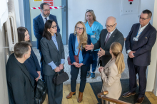 A group of people is standing at the top of a staircase in a building, talking during a visit; one person is making a hand gesture, and next to them are staff members in medical attire.