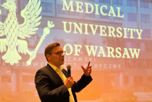 A man in a suit is standing in front of a large screen displaying the university’s name and speaking into a microphone. He raises his other hand to emphasize his statement. He is looking toward the audience.