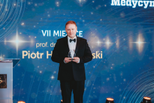 A person in a suit stands on stage next to a large screen with the words “Puls Medycyny” (Pulse of Medicine). The screen displays graphics announcing the first place winner in the poll.