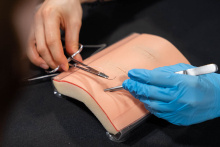 A close-up of surgical suturing practice: two people working on a skin trainer, using instruments such as a needle holder and forceps to perform precise stitches.