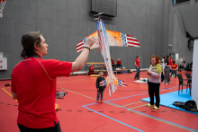 One person is holding a tethered model airplane that is flying in the air. A child is standing opposite and watching the flight with curiosity. Next to them, a woman is holding accessories and looking toward the model. In the background, several people are talking and taking part in activities in the sports hall.