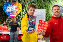 Two young men are standing next to each other and presenting items related to the WOŚP Grand Finale. One is holding a colorful fundraising tin, and the other is holding a sheet of red heart‑shaped stickers. Both are extending their hands toward the camera. In the background, a volunteer station and a large colorful heart with the finale number are visible.
