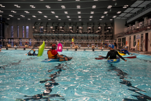 Five people wearing helmets and vests are kayaking in a large indoor pool. The participants maintain their balance and maneuver next to one another. In the distance, several people are watching the activity from the poolside. The water is calm, and the brightly lit hall creates a light background.