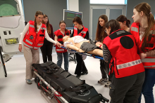 A group of participants in rescue vests carrying a manikin on a stretcher during medical rescue training, next to prepared equipment and a simulat