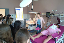 A person conducting a class in a delivery room, presenting the equipment and procedures at a station with a patient manikin lying on a birthing bed, while a group of participants observes the demonstration.