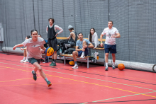Seven people are participating in basketball practice in a sports hall. One person is running to the left while dribbling the ball. Several people are standing or sitting on a bench and watching the exercise. One person is holding a ball, and another is getting ready to enter the court. The participants appear focused.
