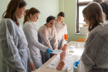 A group of people in protective gowns practicing intravenous access on an artificial limb, observing and discussing each step of the procedure.