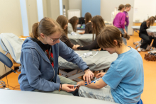 A person practicing the insertion of an intravenous cannula on a partner’s forearm using a training kit, while in the background other participants perform similar tasks at medical stations.