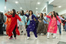 A group of dancers performing an energetic choreography in colorful traditional costumes, dancing in synchrony in the lobby area while an audience gathers around them.