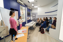 A person leading a session in a museum education room is discussing exhibits and medical models, standing in front of a group of participants seated on benches and listening attentively to the presentation.