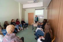 A person leading a class stands by a screen displaying a presentation and discusses educational topics, while a group of participants sits in a semicircle.