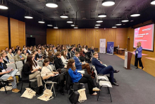 Duża grupa osób siedzi w sali konferencyjnej i słucha prelegentki mówiącej przy mównicy. Na ekranie obok znajduje się slajd prezentacji.