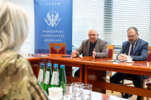 Three people are sitting at a table in a conference room. Two of them are on the right side of the table, and one is on the left. The person on the left is wearing a military-style uniform, while the people on the right are dressed in elegant clothing. One of them is gesturing with their hands, and the other has their hands placed on the table. In the background, there is a blue board with the inscription 'Medical University of Warsaw'.