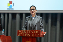 A woman is standing behind a lectern with the inscription 'Veritatis Splendor' and speaking into two microphones. She is resting one hand on the lectern while making a gesture with the other beside it. Next to her, there is a monitor placed on a stand. In the background, part of a screen with graphic elements is visible.