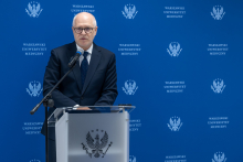 A person is standing behind a lectern and speaking to the audience. They are resting their hands on the podium and looking ahead. A microphone is positioned right next to them. In the background, a blue wall with the repeating logo of the Medical University of Warsaw is visible.