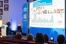 A speaker stands at the lectern, delivering a presentation. Next to them is a tall screen displaying a chart and photos of several people. Four individuals are seated in the audience, looking toward the stage. The lighting focuses attention on both the screen and the speaker.