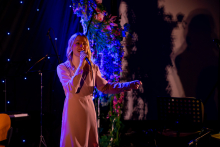 A woman is standing on stage and singing into a microphone. She is holding the microphone with one hand and making a gesture toward the audience with the other. Her posture is relaxed, and her facial expression suggests focus. Behind her, colorful lighting and stage decorations are visible.