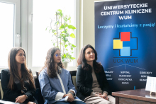 Three individuals are seated next to each other on chairs, with a banner of the University Clinical Center of WUM featuring a colorful logo and the slogan ‘We treat and educate with passion!’ in the background. On the left, a window with a climbing plant is visible.