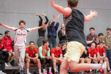 A player is running with outstretched arms across the sports hall, while the team and the fans on the bench and in the stands react enthusiastically, raising their hands and clapping.