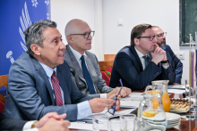 Three people are sitting at a table in a conference room. They are dressed elegantly and appear to be listening thoughtfully. On the table there are documents, pens, and glasses of water. In the background on the left, a blue banner with white symbols and partially visible text can be seen.