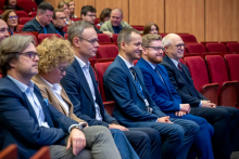 People are seated in several rows of red chairs in an auditorium, dressed in formal attire, listening to an event or lecture.