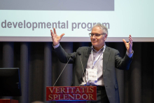 A person speaking at a podium, gesturing broadly during a presentation, with part of a slide containing text visible in the background.