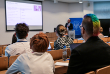 A group of people sitting in a lecture hall. In front of them is a multimedia screen displaying a presentation. On the right side, a man holding a microphone is standing.