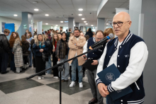 A person standing by a microphone in a lobby area, holding a folder with documents, against the background of a group of participants waiting in line during the event.