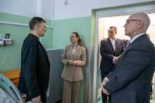Several people are standing and talking in a small room with green and white walls; one person on the left is leading the conversation, while the others listen, standing by the door and next to a cabinet.