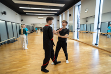 Two people practicing self‑defense techniques in a training room, standing opposite each other in a preparatory stance; mirrors and other class participants are visible in the background.