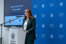 A person is standing at a lectern and delivering a speech. They are looking toward the audience and resting their hands on the podium. A microphone is positioned next to the lectern. Beside it, there is a blue wall featuring the logo of the Medical University of Warsaw. On the screen nearby, a slide appears with a title concerning the health of the residents of Warsaw.