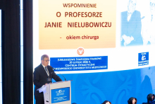 A speaker stands at the lectern, delivering a presentation during the symposium. He rests his hands on the podium and looks toward the audience. Behind him is a large screen displaying the title of the presentation dedicated to the memory of Professor Jan Nielubowicz, along with archival graphics. At the bottom of the screen, information about the jubilee event is visible.