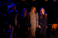 Four women are standing next to each other on stage and singing.