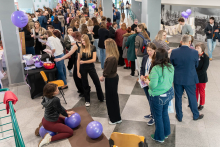 An event with numerous attendees is taking place in the lobby area. Conversations at the booths, exercises on mats using purple balloons, and tables with informational materials can be seen. A crowd of participants fills the entire hall.