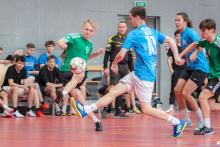 Players in green and blue jerseys are fighting for the ball during a dynamic action in an indoor football match, while the remaining participants watch the situation from the bench