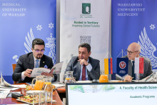 Three elegantly dressed men are seated at a table. The man on the left is looking through an information folder he is holding in his hands. The other men are looking in his direction. On the table there are documents and small institutional flags. In the background, two banners are visible: on the left, ‘Medical University of Warsaw,’ and on the right, ‘Universidad del Quindío.’