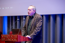 A person standing at a lectern with the inscription ‘Veritatis Splendor’, speaking into two microphones in an auditorium. In the background, curtains and a portion of a projection screen are visible.