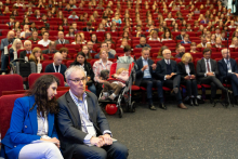 Audience seated in a large lecture hall; in the foreground two people sitting in the front row, with many attendees filling the red seats behind them.