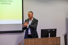 A formally dressed man is standing by a white wall, holding a microphone in his hand. He is speaking. Next to him, a fragment of a multimedia screen displaying a presentation is visible.