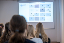 Grupa uczestników siedzi w sali i patrzy na ekran z prezentacją zawierającą piktogramy i nazwy kierunków studiów medycznych. Ekran jest głównym elementem kadru.