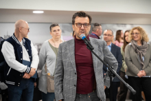 A person speaking into a microphone during an indoor event, surrounded by gathered participants standing in the background.