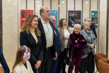 A group of people is standing in a hall with light, marble walls, looking in the same direction; behind them, colorful posters of cultural events are visible on the walls.