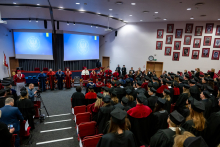 Several hundred PhD holders and habilitated doctors in academic gowns are standing in the auditorium, facing the university authorities. In front of them stands a group of professors in ceremonial gowns. Behind the lectern stands the vice‑rector, with other members of the university leadership seated nearby