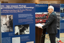 A speaker at a lectern during an event accompanying the exhibition on the history of clandestine teaching at the Faculty of Pharmacy of the University of Warsaw, set against display panels featuring descriptions and archival photographs.