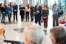 Grupa osób stoi w półkolu podczas wydarzenia w przestrzeni wewnętrznej. Na pierwszym planie widoczne są dwie postacie stojące za mikrofonem, a w tle widać uczestników oraz fragment stołów i ekspozycji.