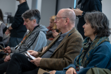 Several people are sitting in rows and participating in a meeting. The person in the foreground is holding a notebook and looking toward the speaker. Next to them sits another person facing the same direction. In the background, more people can be seen following the event. A few individuals are standing at the back of the room operating equipment.