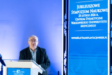 A speaker stands at the lectern, delivering a presentation during the symposium. He rests his hands on the podium and looks toward the audience. Next to him is a tall screen displaying information about the jubilee event.