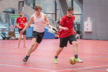 Two players are competing for the ball during an indoor football match, running close to each other across the pitch, while the other players watch the action in the background.