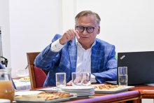 A man sitting at a table. He is dressed elegantly and wearing glasses. He is gesturing with one hand, which is raised in the air.