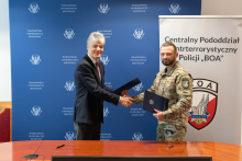 Two men are standing next to each other and shaking hands. One is holding a black folder, while the other is holding a navy-blue folder with an emblem. The person on the left is wearing an elegant outfit, and the person on the right is dressed in a military-style uniform. In the background, there is a blue board with the inscription 'Medical University of Warsaw' and a white banner with the logo and the inscription 'Central Counterterrorism Subunit of the Police BOA'.